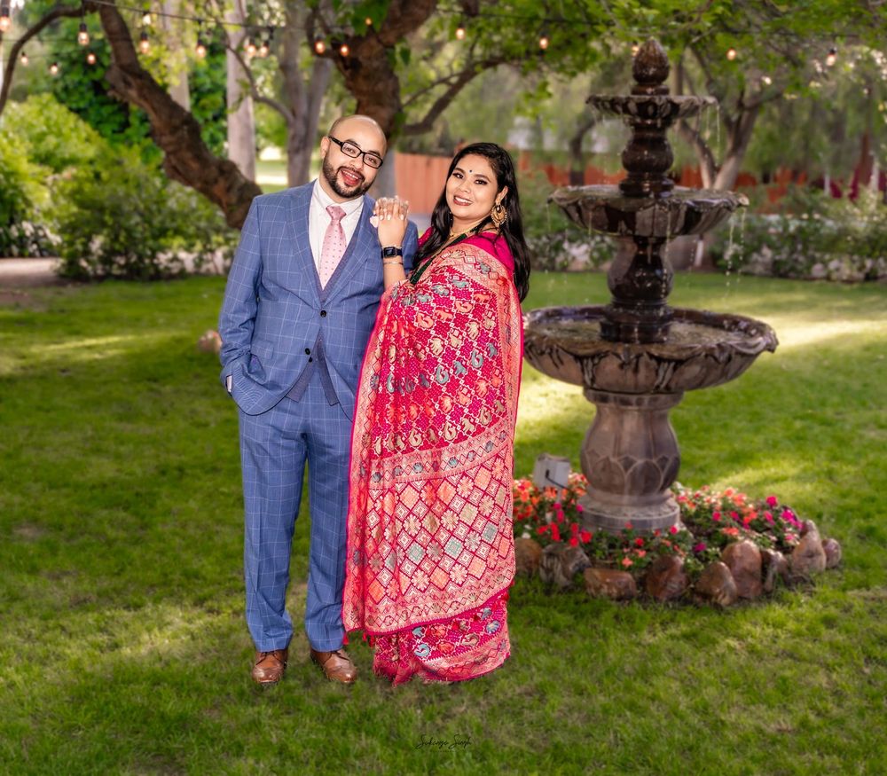 Couple posing by stone fountain in garden, man in blue suit, woman in red embroidered sari, lush green trees background.