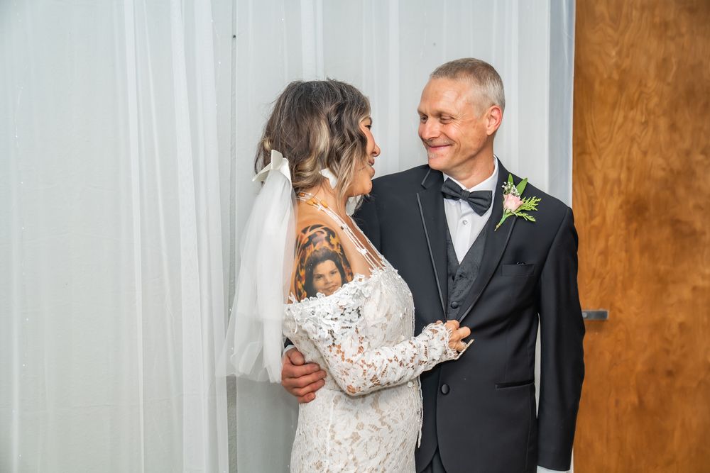 Bride with tattoos in lace wedding dress embraces smiling older man in tuxedo at wedding ceremony.