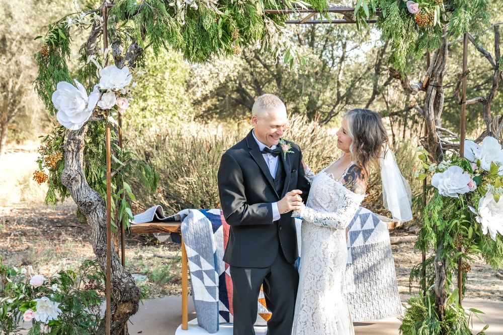 Couple exchanging vows under floral arch with white blooms and greenery in outdoor woodland wedding ceremony.
