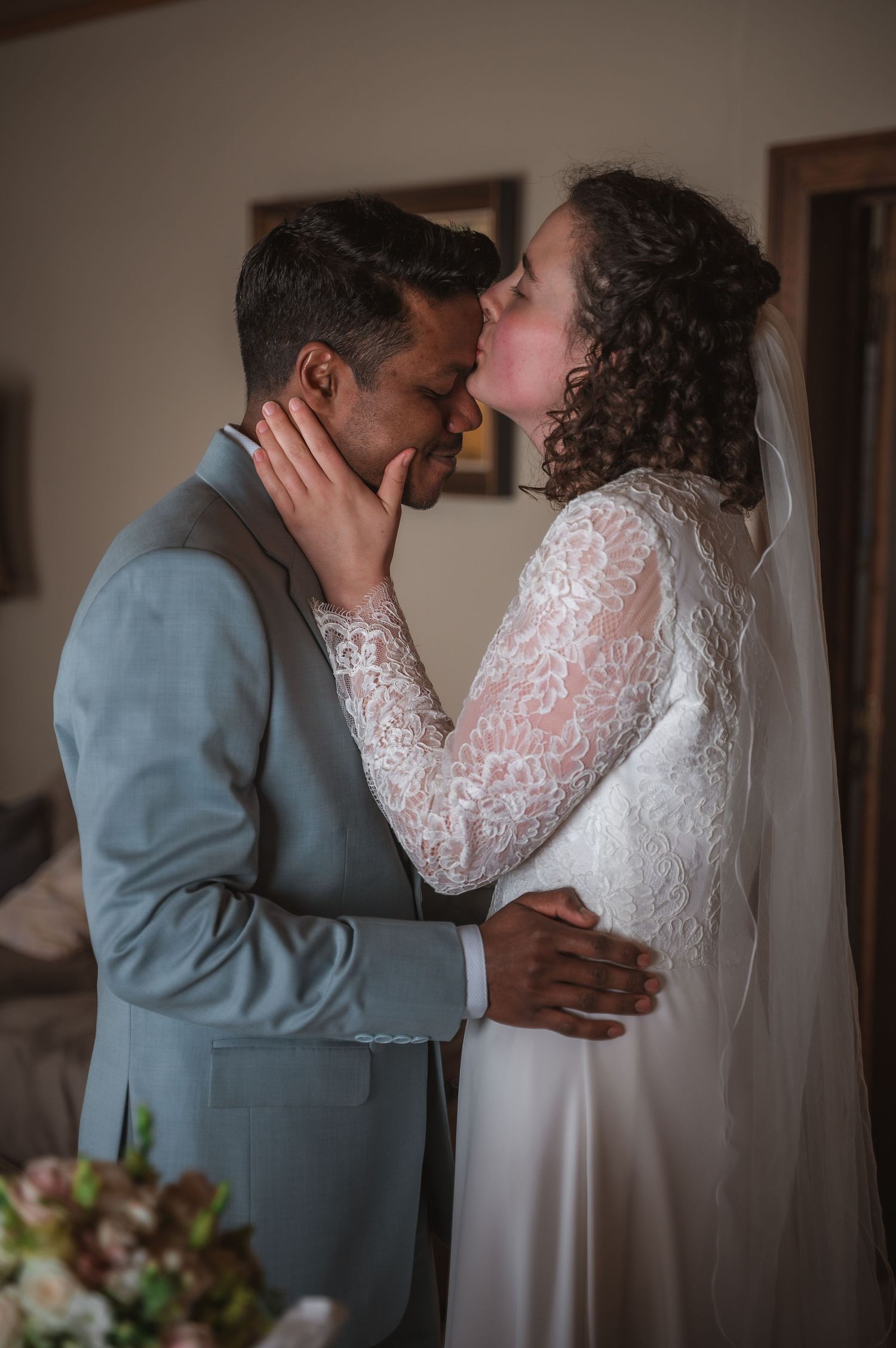 A bride and groom share their first kiss, dressed in a white lace gown and gray suit, in a warmly lit indoor setting.