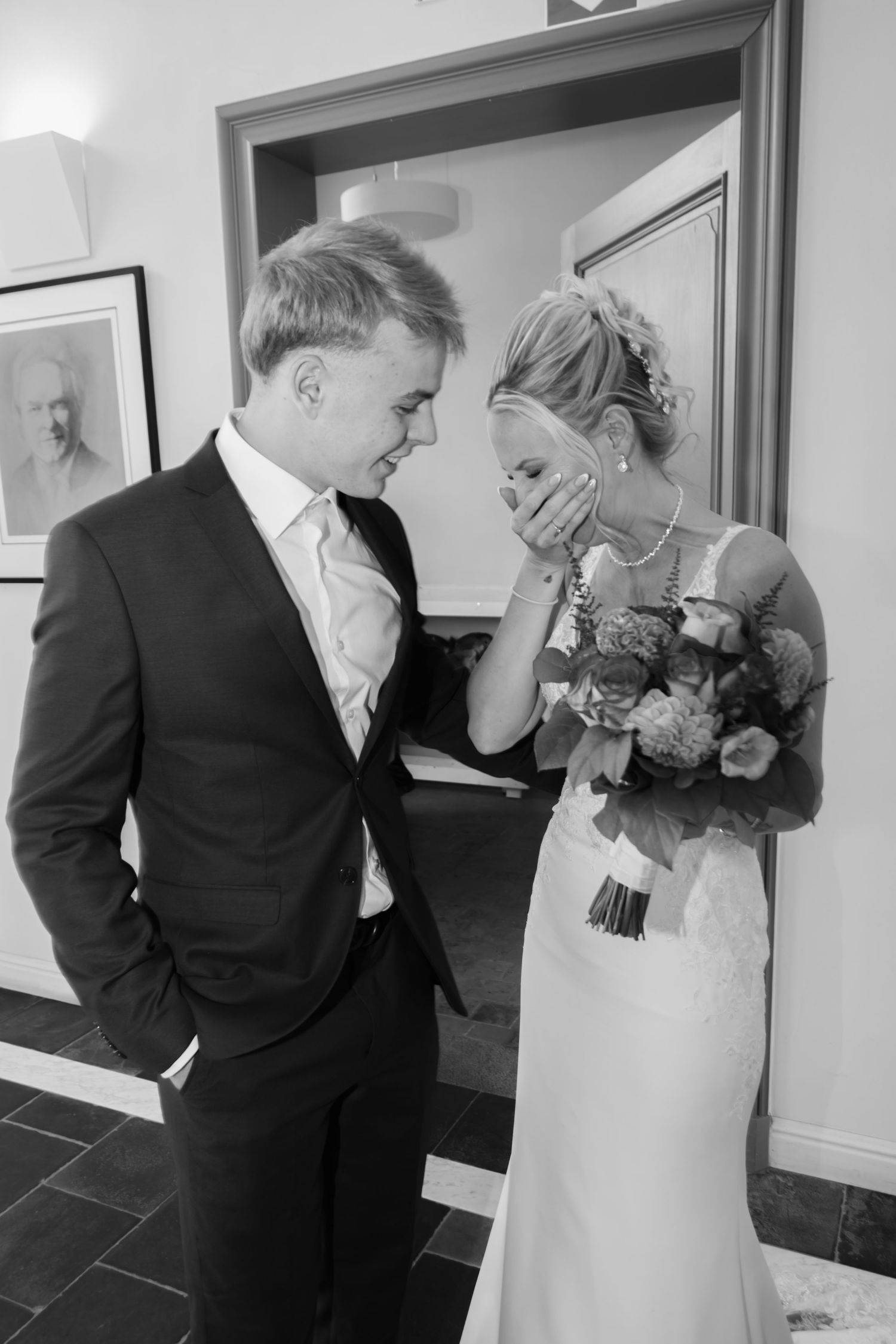 Black and white photo of an emotional bride covering her mouth while holding bouquet, standing with groom in suits.