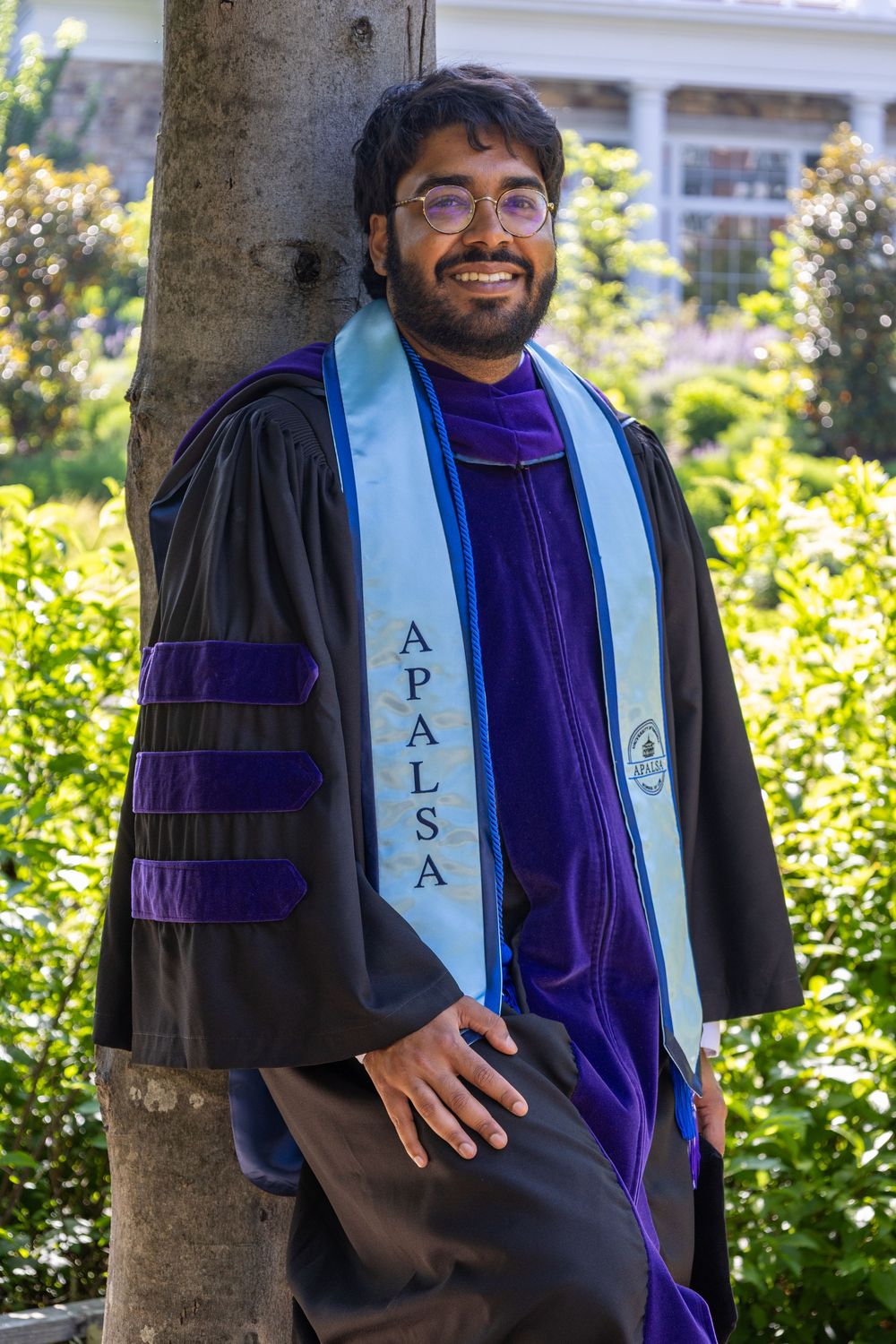 Smiling graduate in black and purple doctoral robes wearing APALSA stole, leaning against tree in garden setting.