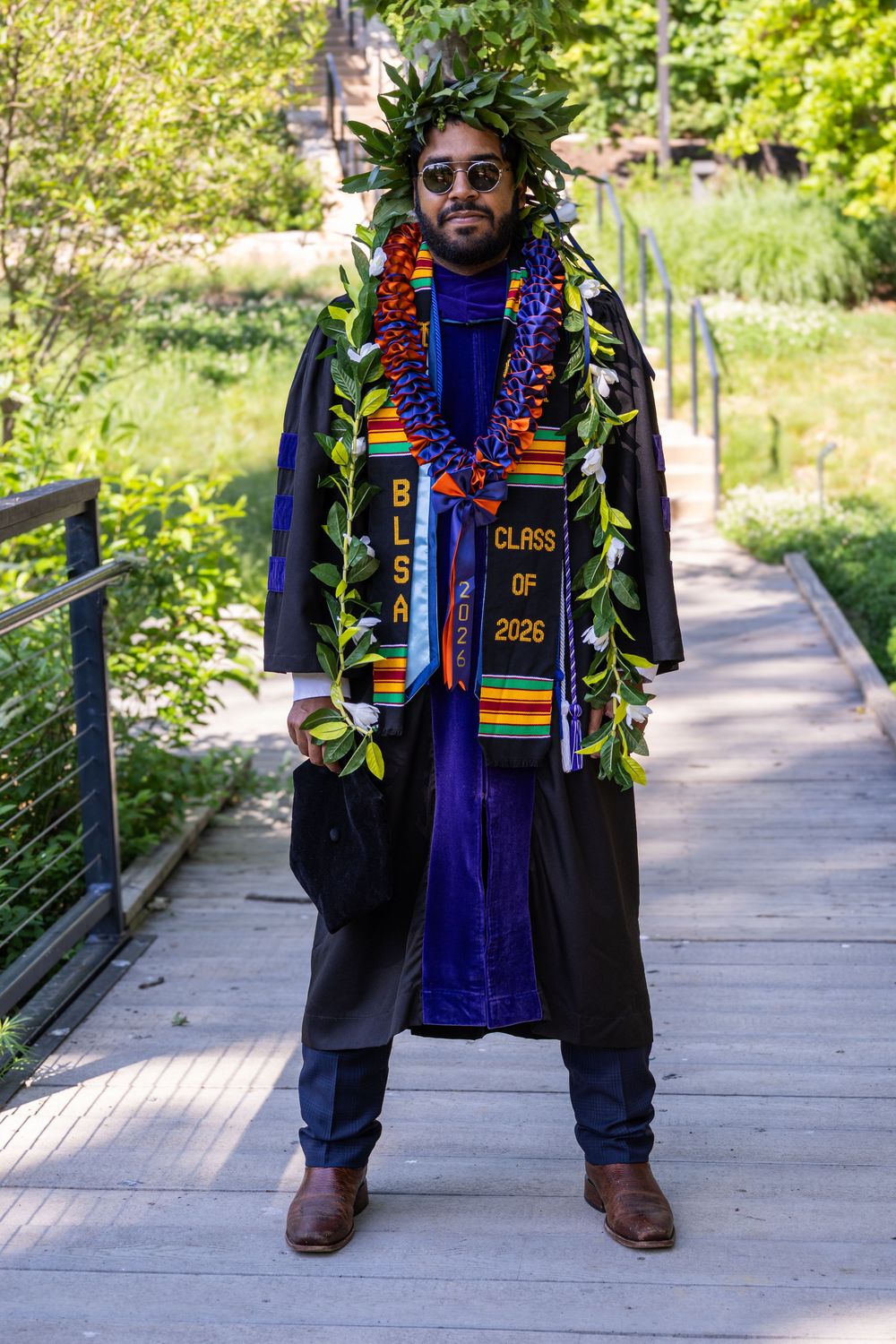 Graduate in black gown with lei, kente stole, and laurel wreath poses on wooden bridge, Class of 2026.