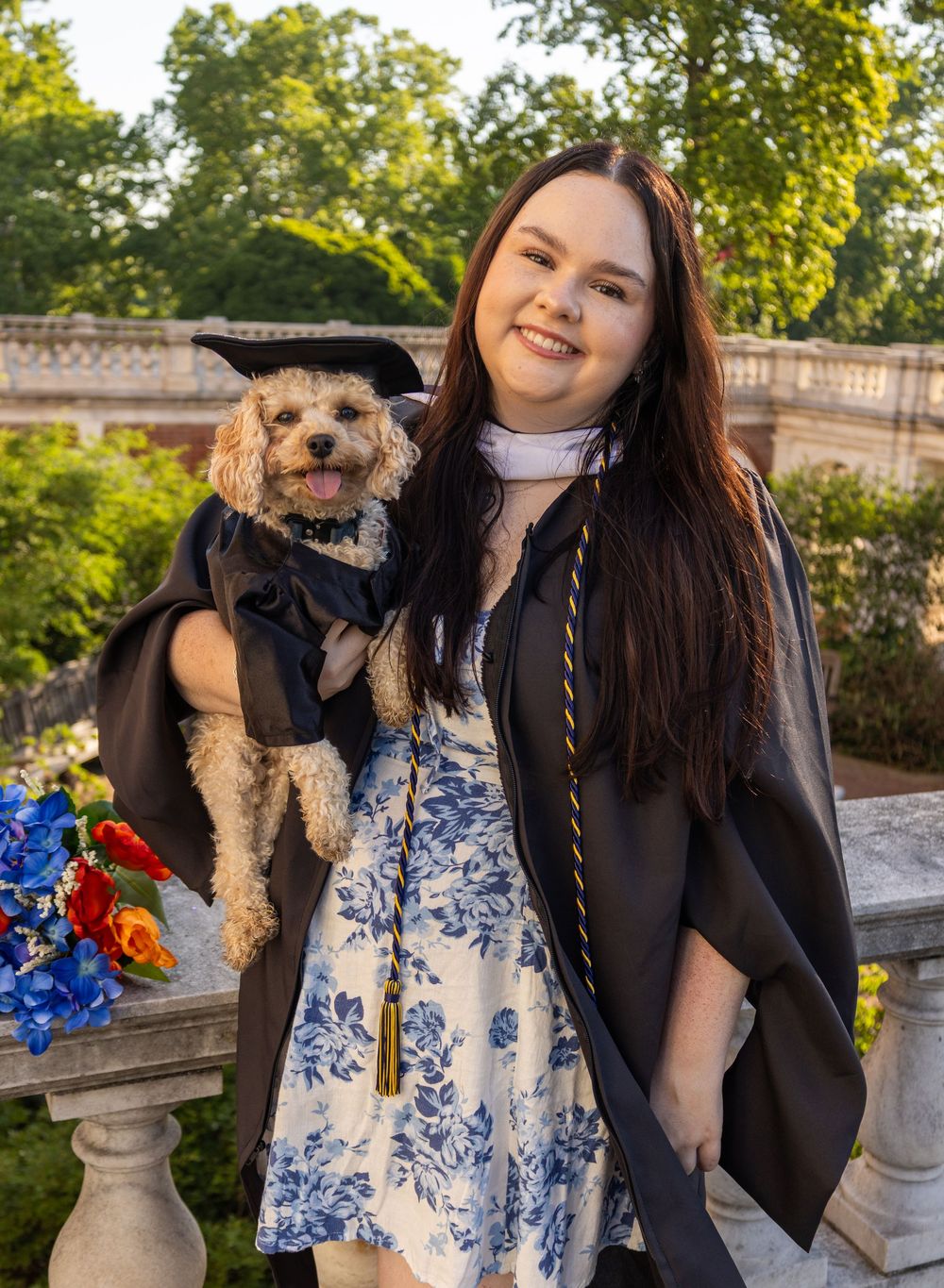 Young woman in graduation gown holds a small dog also dressed in graduation attire, posing outdoors.