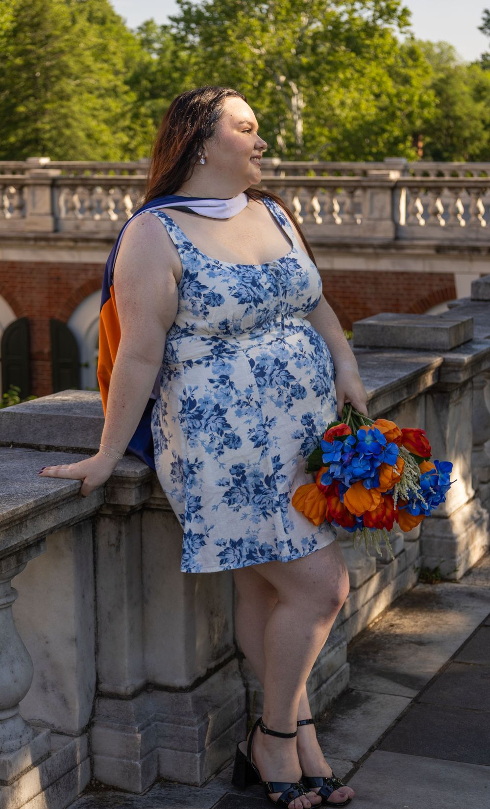 Woman in blue floral dress holding colorful bouquet of flowers, leaning on stone balustrade outdoors.
