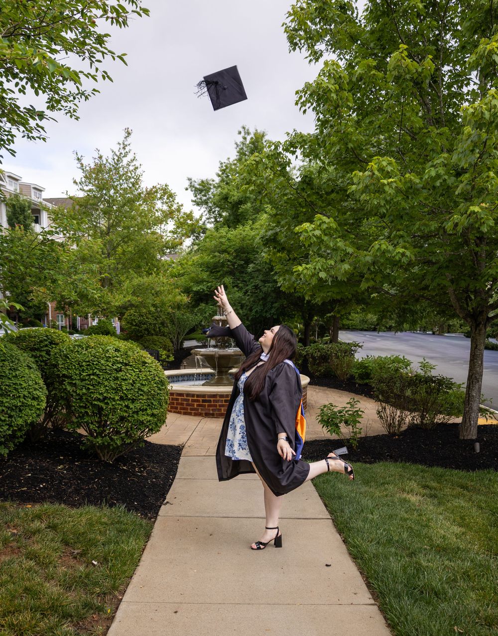Graduate in cap and gown tosses mortarboard into the air on a tree-lined walkway, celebrating academic achievement.