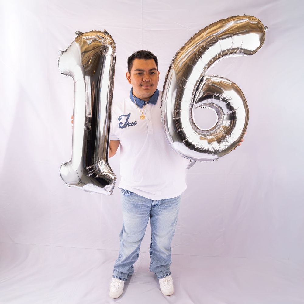 Young man holding large silver number 16 balloons, wearing white polo shirt and jeans, celebrating 16th birthday.