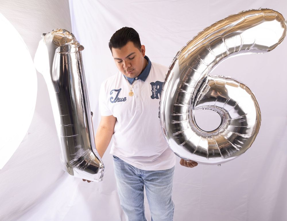Man holding large silver number 16 foil balloons against white background, wearing white polo shirt and jeans.