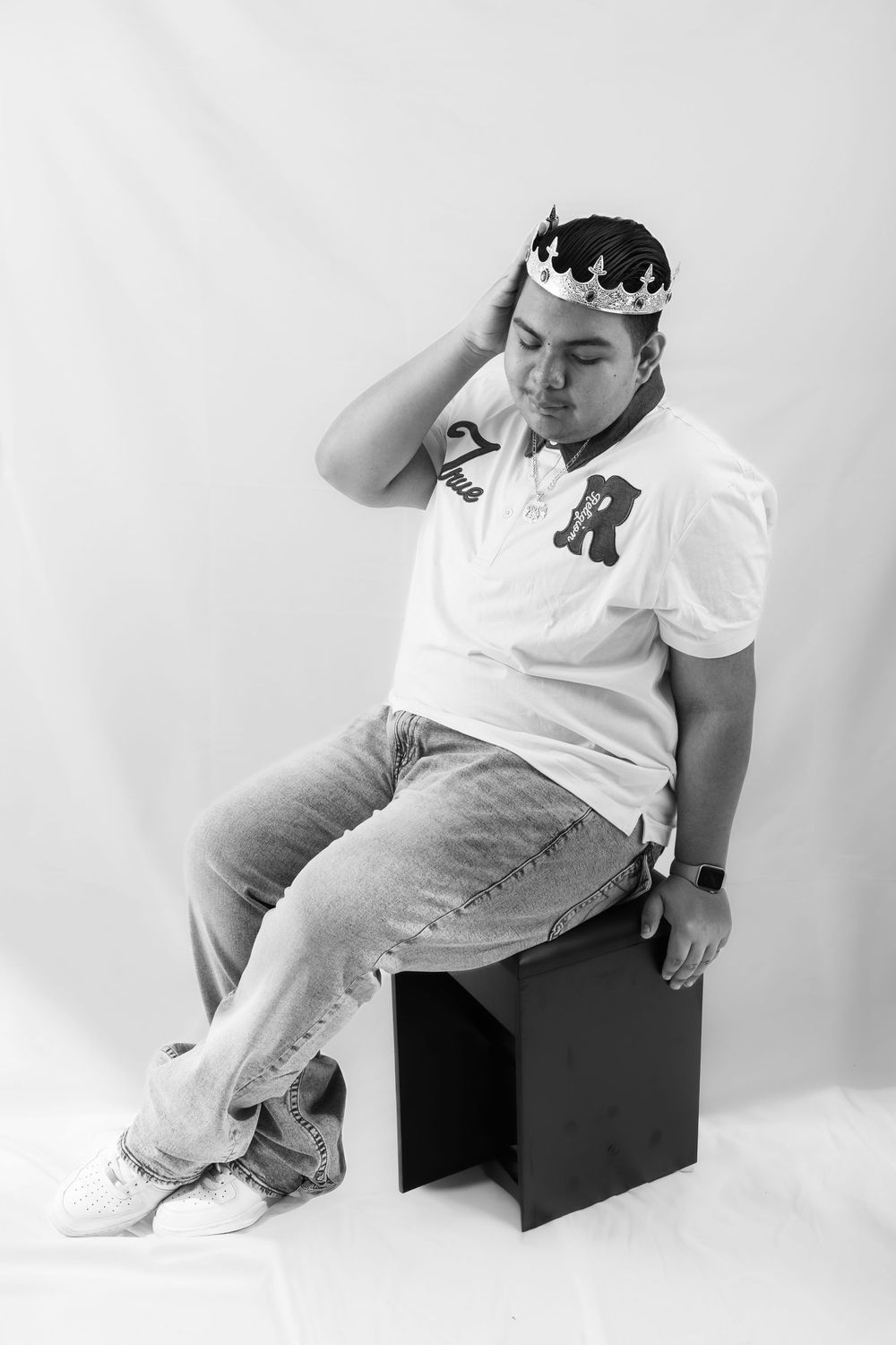 Black and white photo of a young man wearing a crown, white polo shirt, jeans, sitting on a black stool.