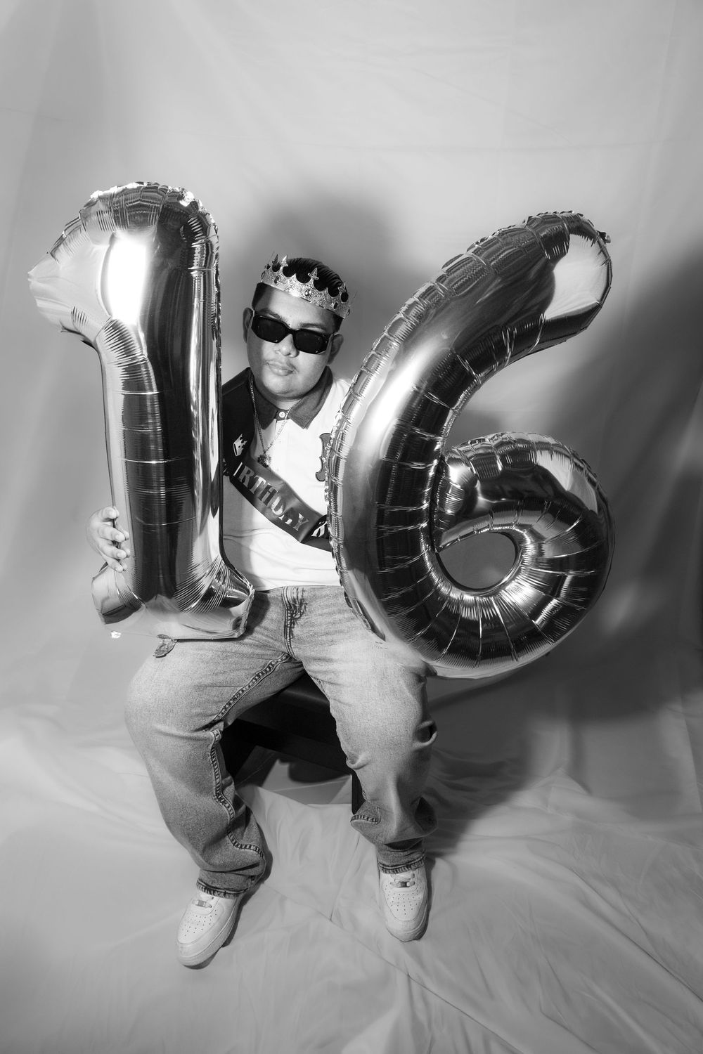 Person holding large silver number 16 balloons wearing crown and sunglasses at a sweet sixteen birthday celebration.