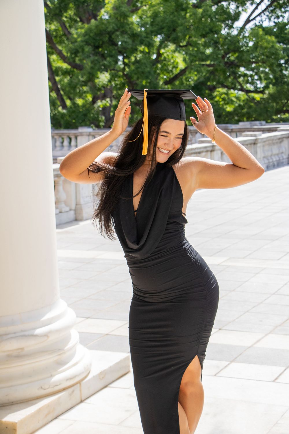 Smiling young woman in a black dress adjusting her graduation cap beside a white column outdoors.