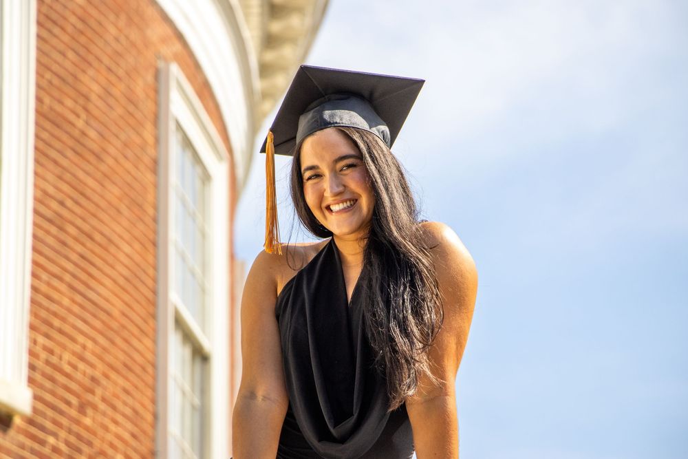 Happy young woman in black graduation cap smiling in front of a brick building on a bright day.
