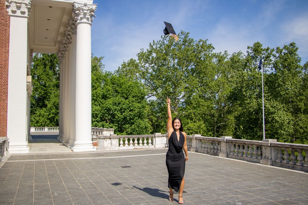 Graduate in black dress tossing her cap in the air near white columns on a sunny campus plaza.