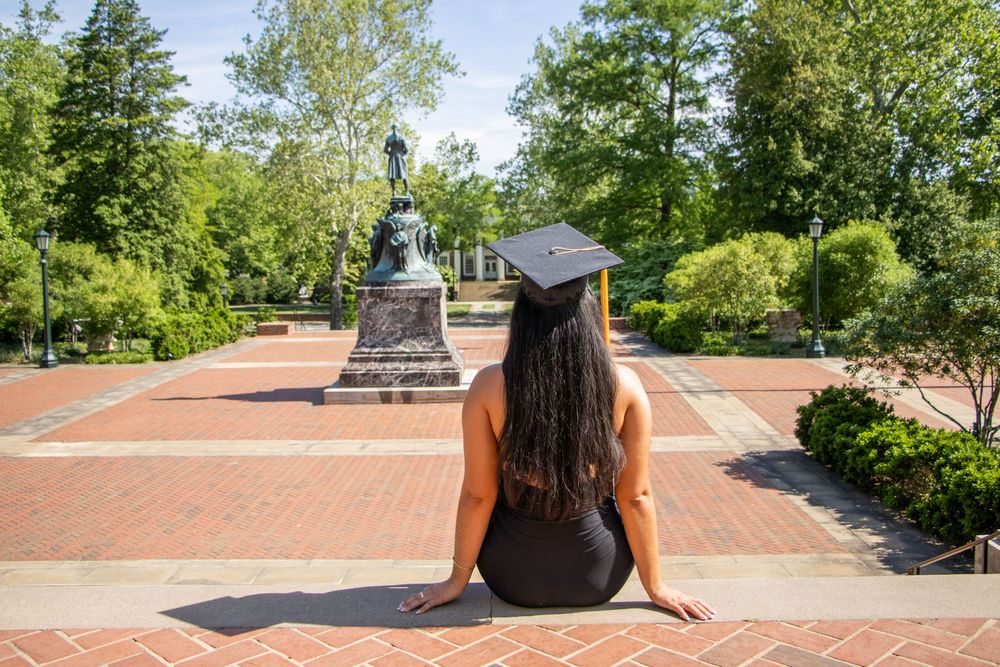 Graduate in black cap sitting on brick steps facing a campus statue surrounded by lush greenery.