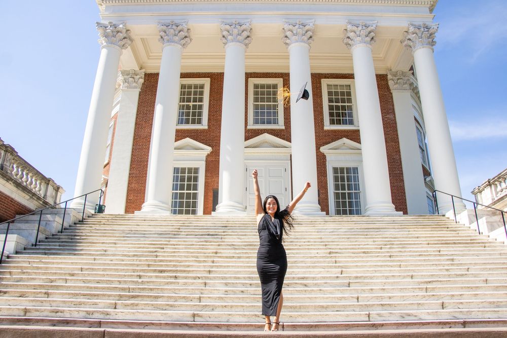 Graduate in black dress throwing cap in front of a grand white-columned brick university building.