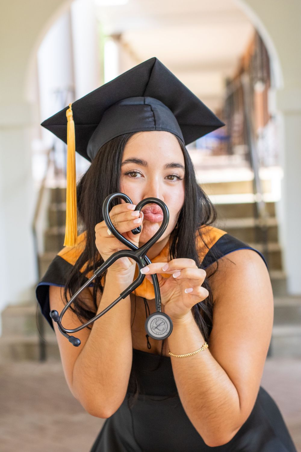 Female graduate in cap and gown forming a heart shape with a stethoscope while smiling indoors.
