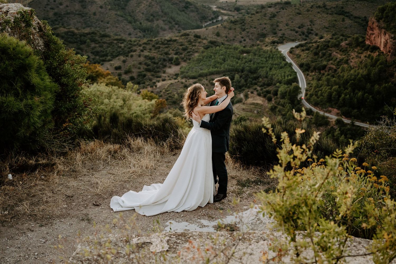 Couple in wedding attire embracing on mountain cliff with winding road visible in valley below.