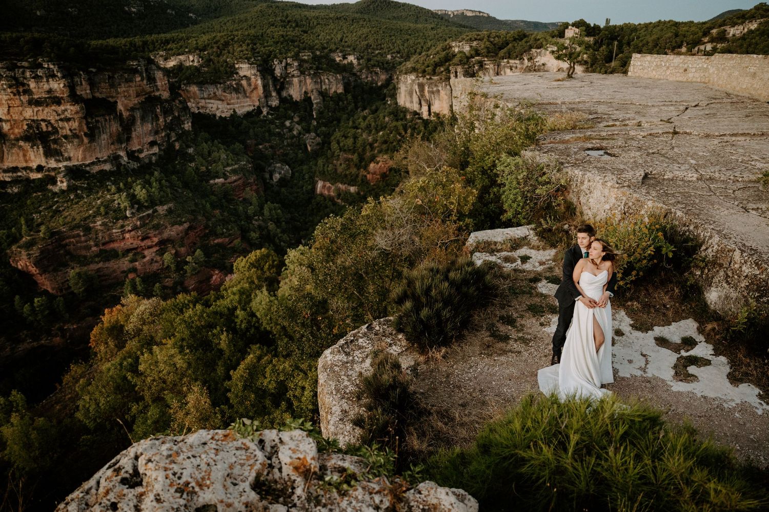 Wedding couple poses on dramatic cliffside overlook with mountain landscape in background.