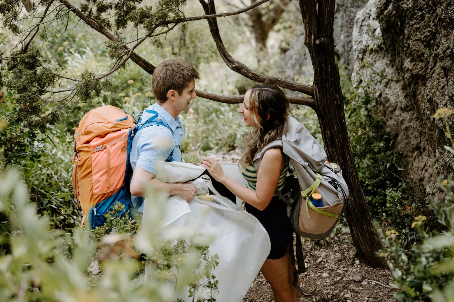 Two hikers with backpacks share a moment while taking a break on a forested trail.
