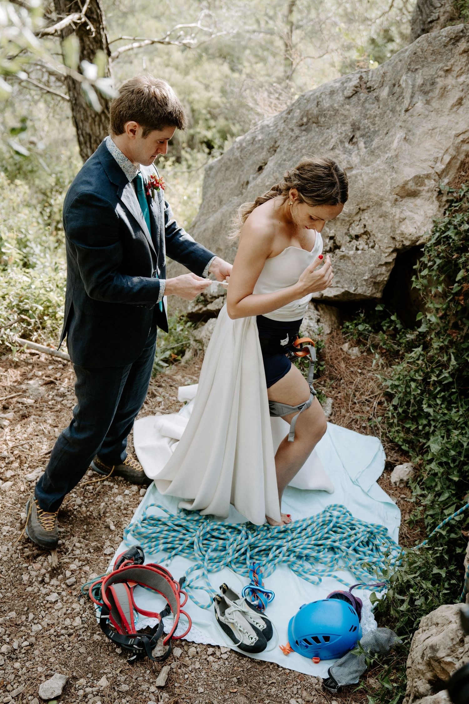 A couple in wedding attire prepares their rock climbing gear on a blue blanket near a cliff face.