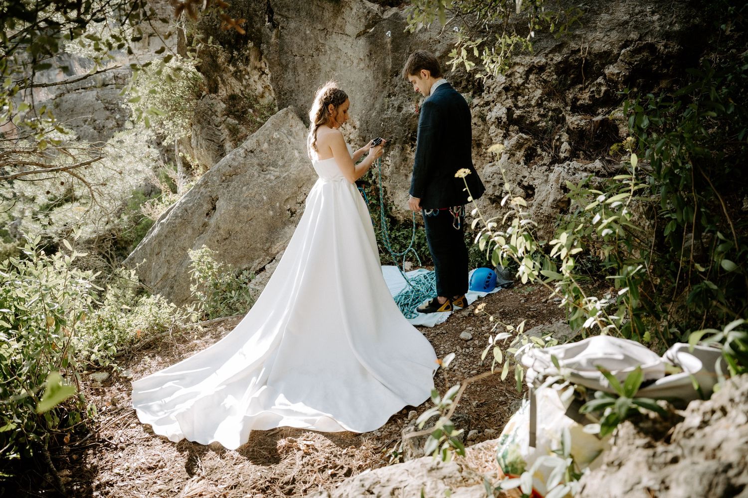 A couple in wedding attire walks along a rocky mountain path with flowing white dress trailing behind.
