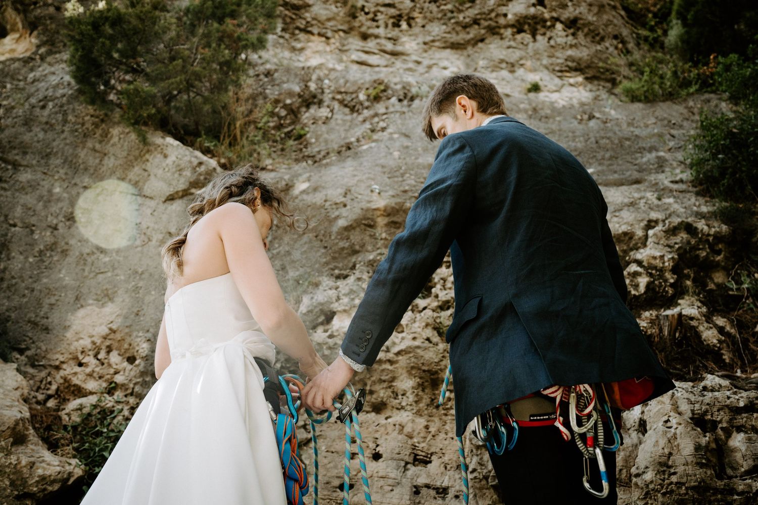 A couple in wedding attire holding hands while rock climbing with safety gear against a rocky cliff face.