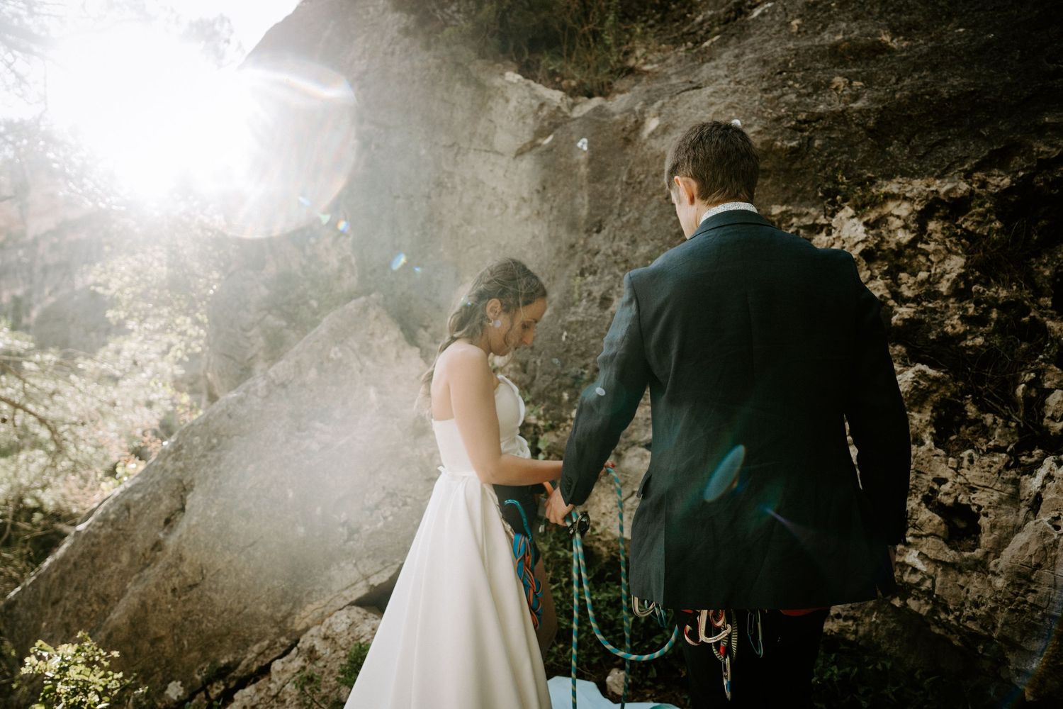 A couple in wedding attire standing near rocky cliffs with sunlight streaming through.