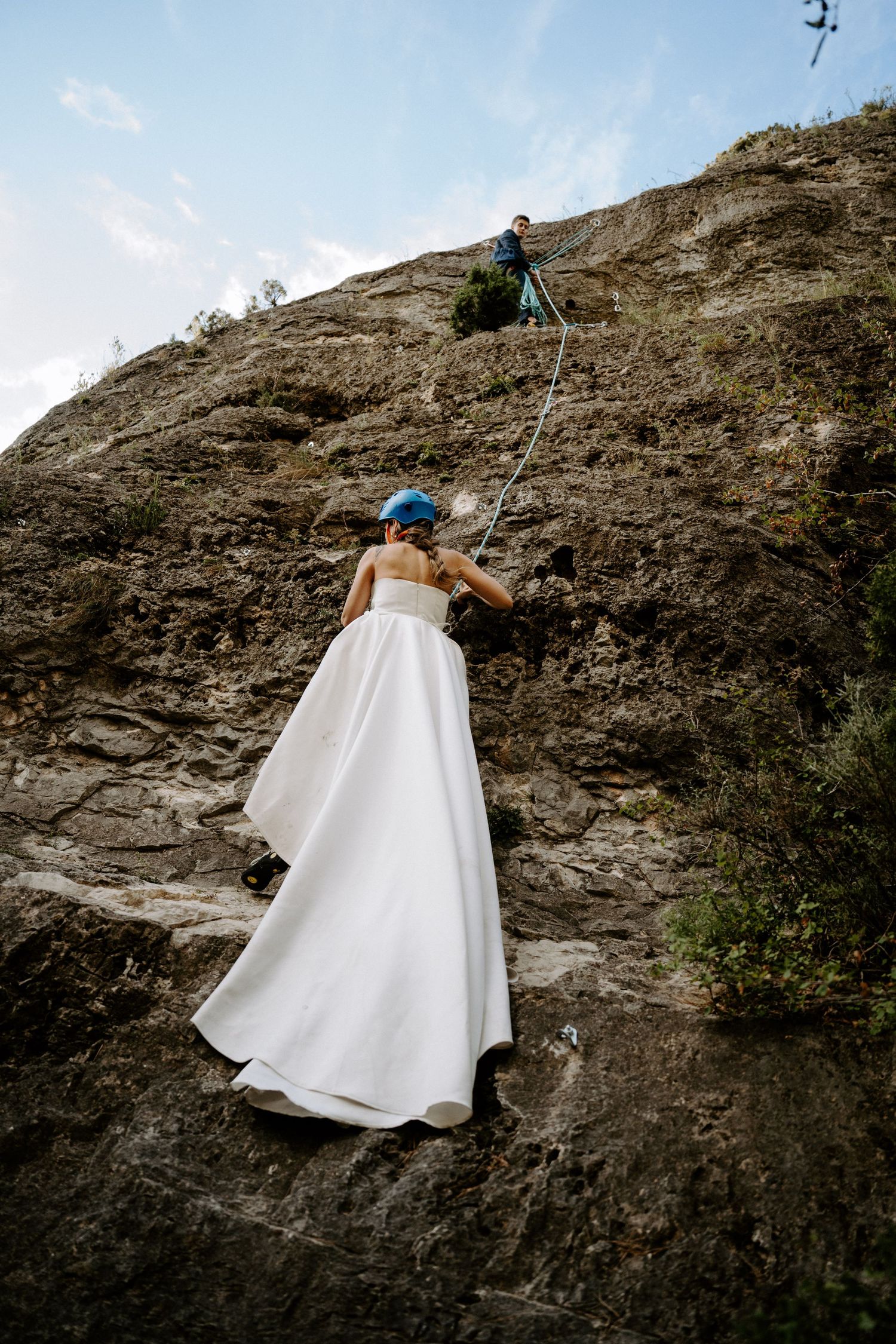 Rock climber in a flowing white dress scaling a steep cliff face with climbing gear.