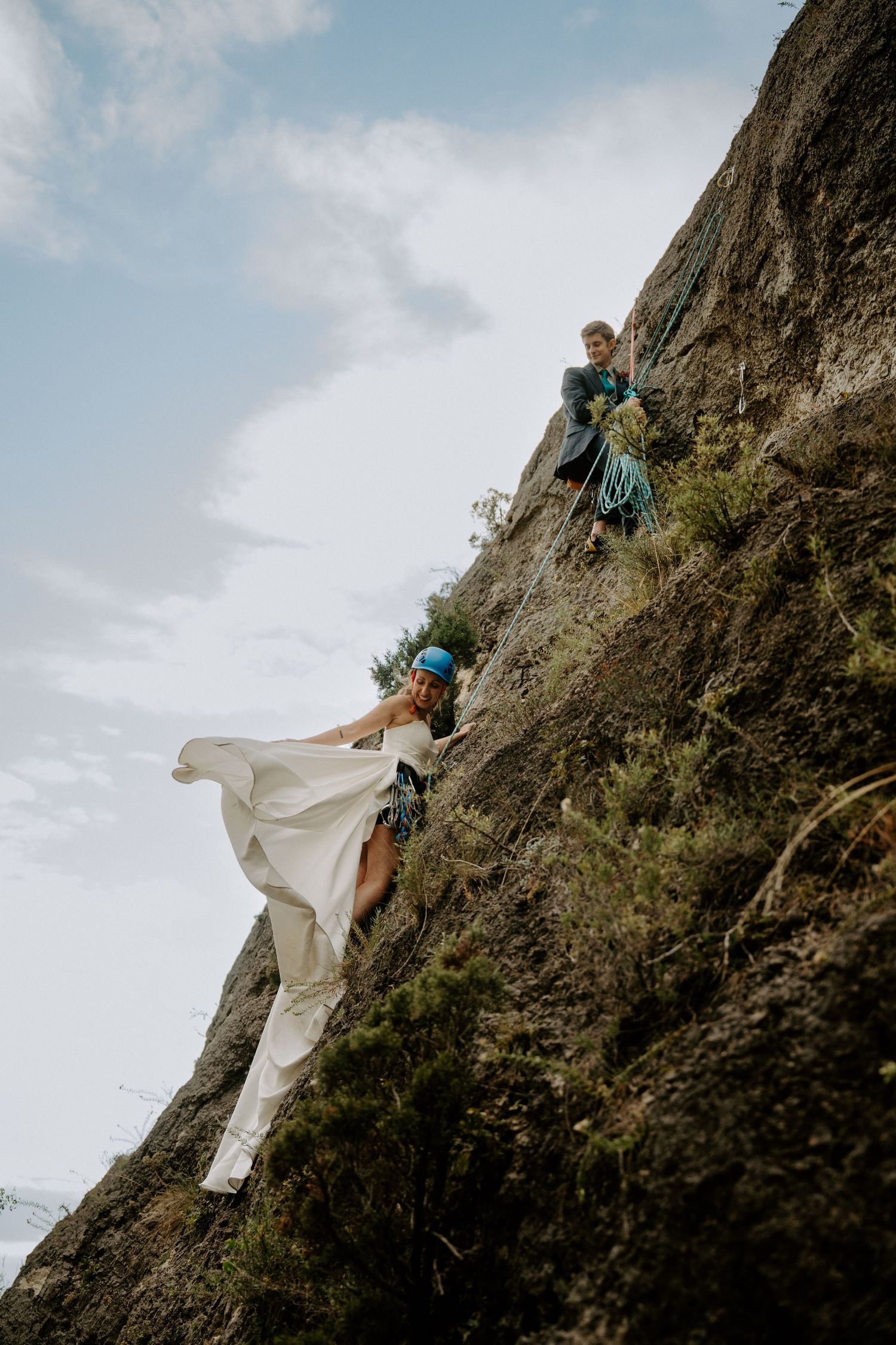 A couple rock climbing in wedding attire scales a steep cliff face against a cloudy sky.
