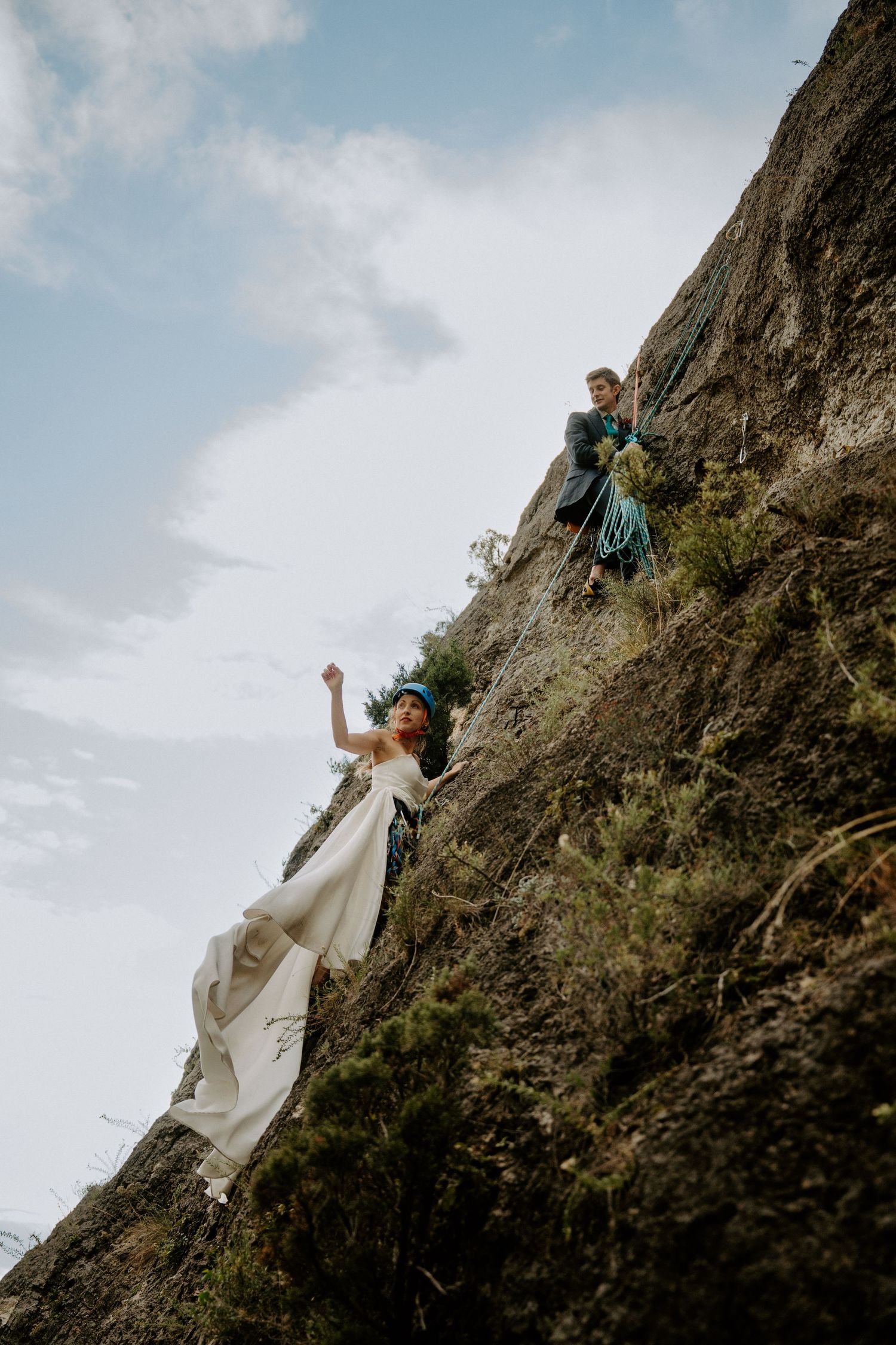 Two people in formal attire rock climbing on a steep cliff face against a blue sky.