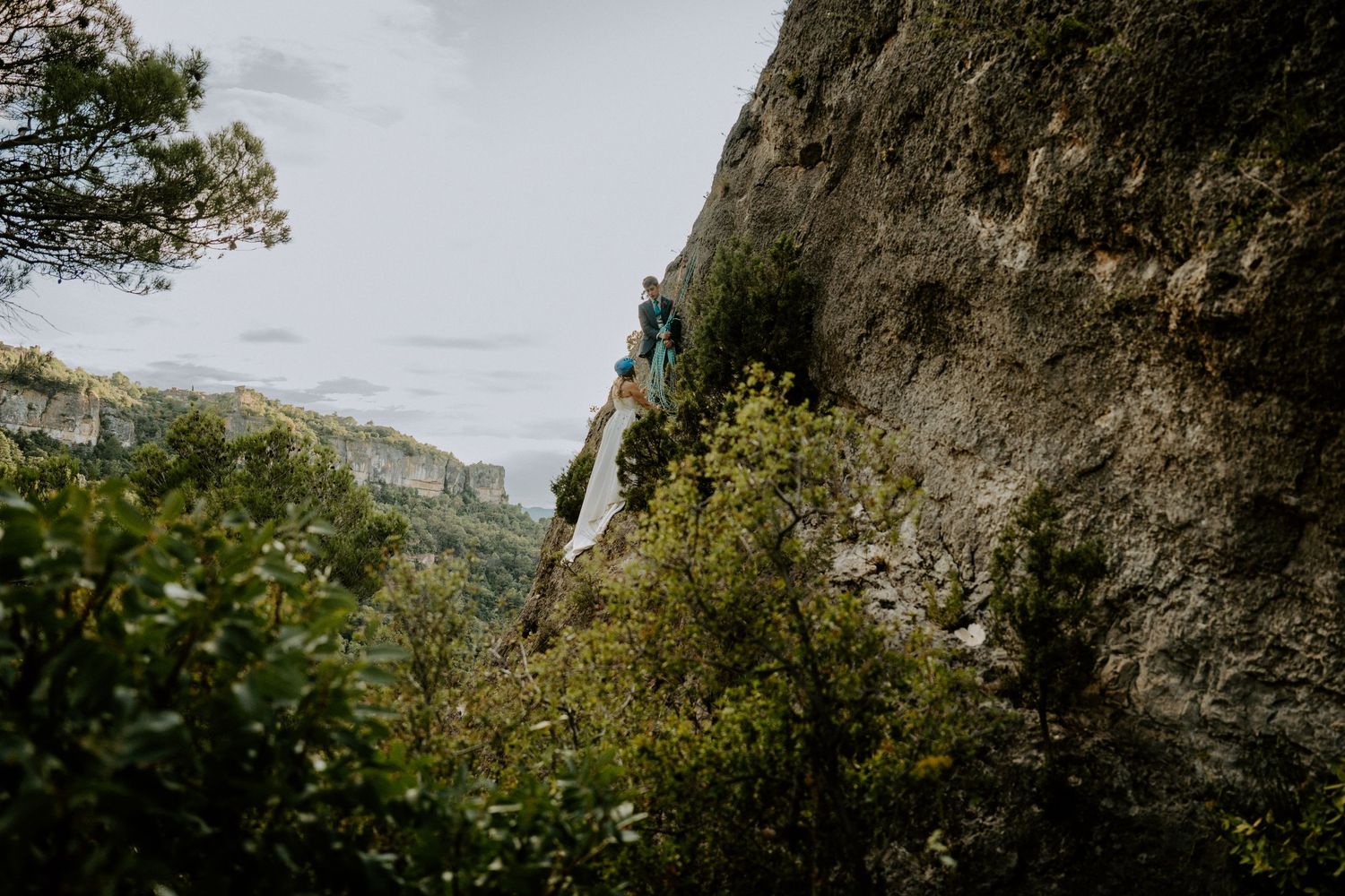 A climber scales a tall limestone cliff face against a cloudy mountain backdrop.