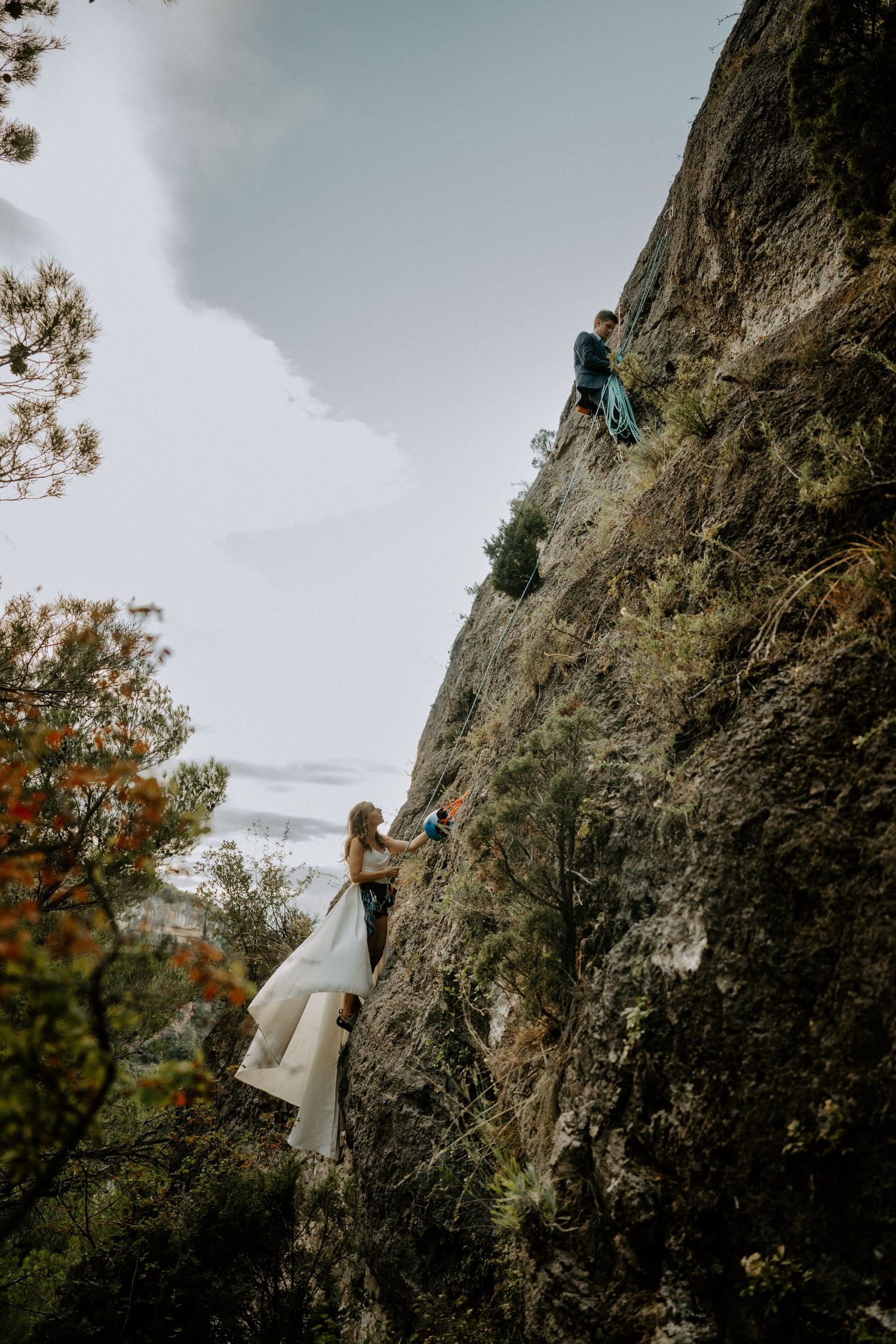 Rock climbers scale a steep cliff face with one person in a flowing white dress during an outdoor adventure session.