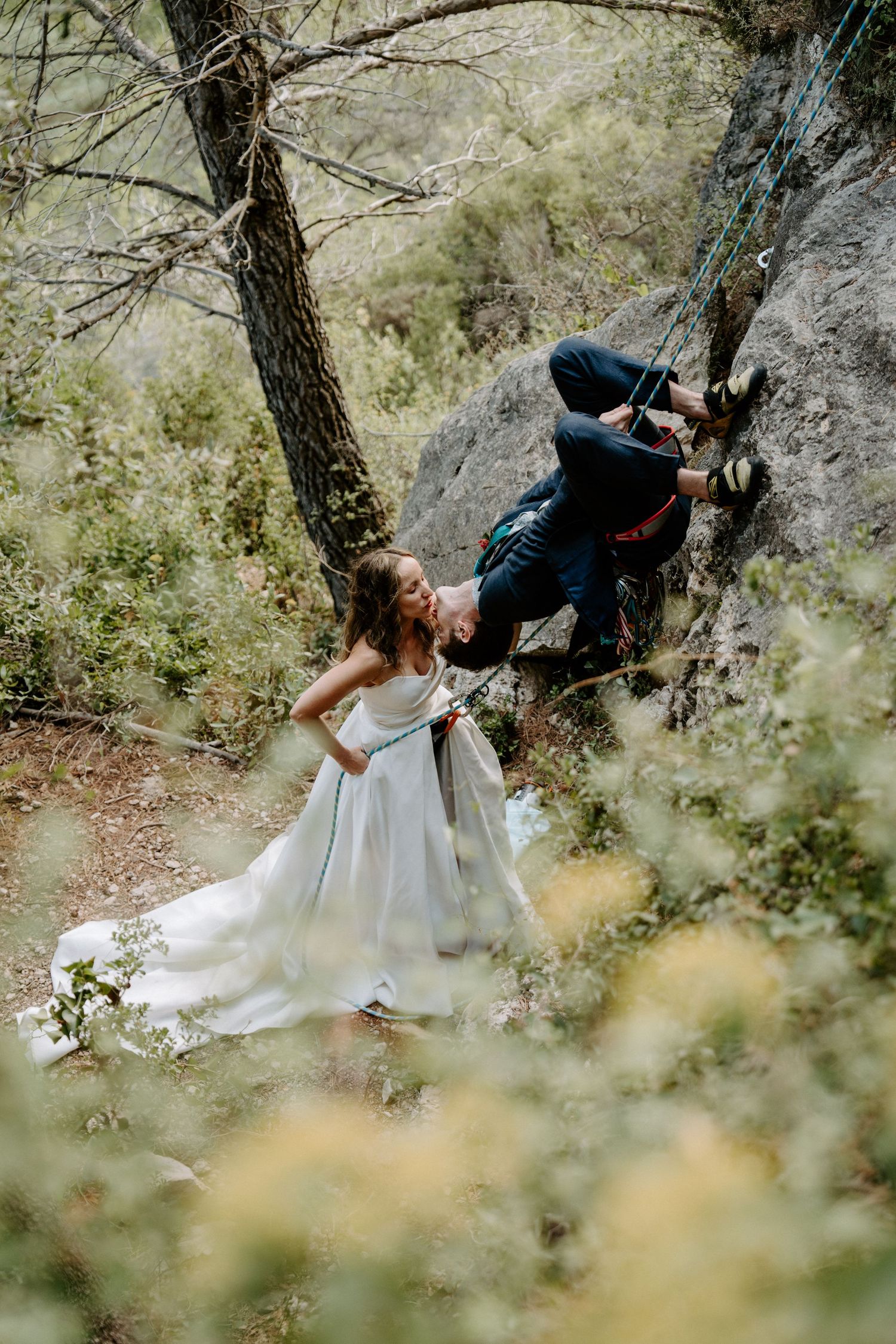 Wedding couple performs adventurous rock climbing moves through trees and rocky terrain.