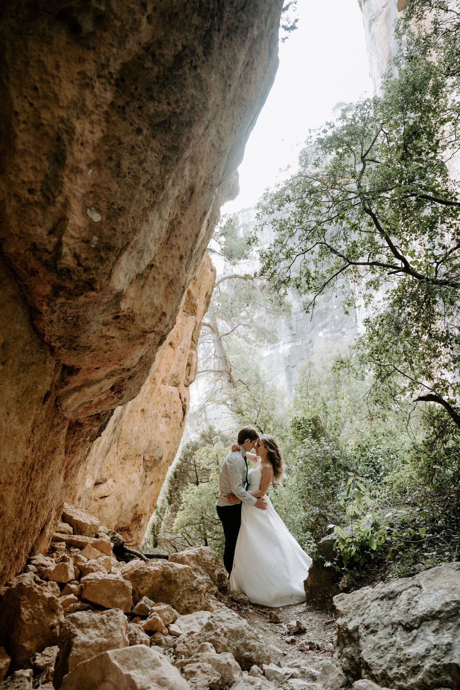 A couple in wedding attire share a romantic moment under a dramatic rock formation in a misty forest setting.