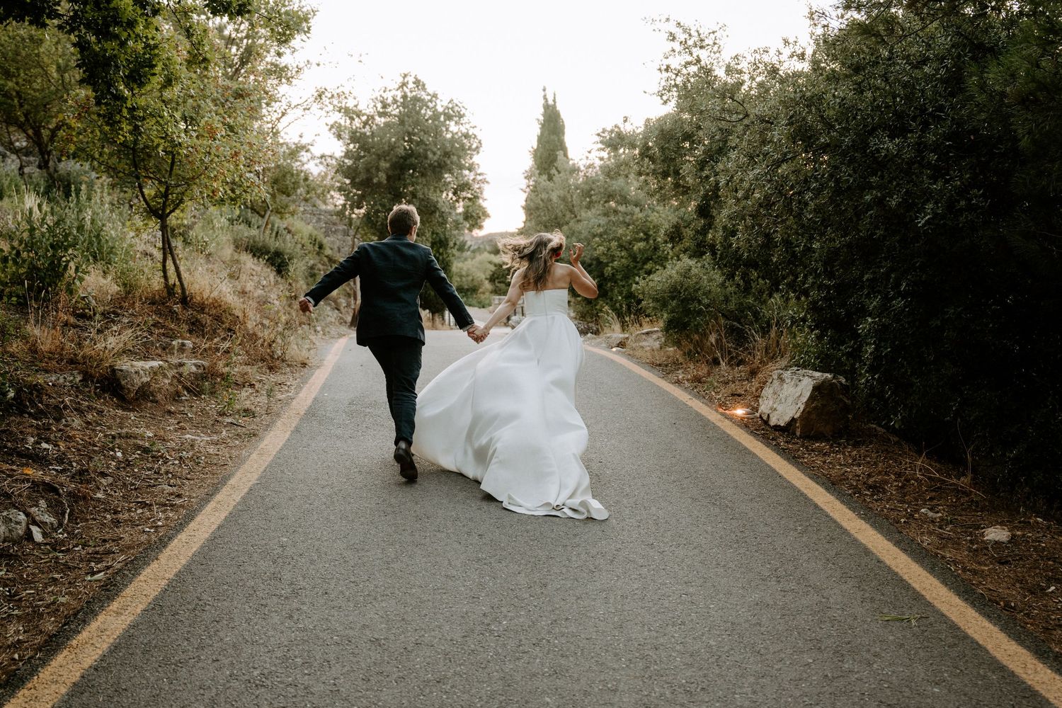 A couple in wedding attire running together on a paved road surrounded by trees.