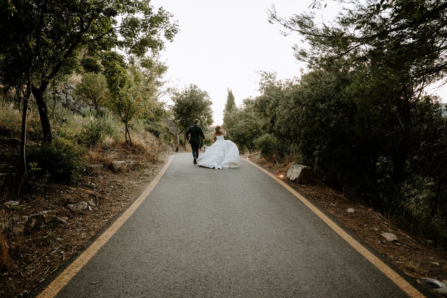 White wedding dress flowing on a tree-lined paved road leading into the distance.