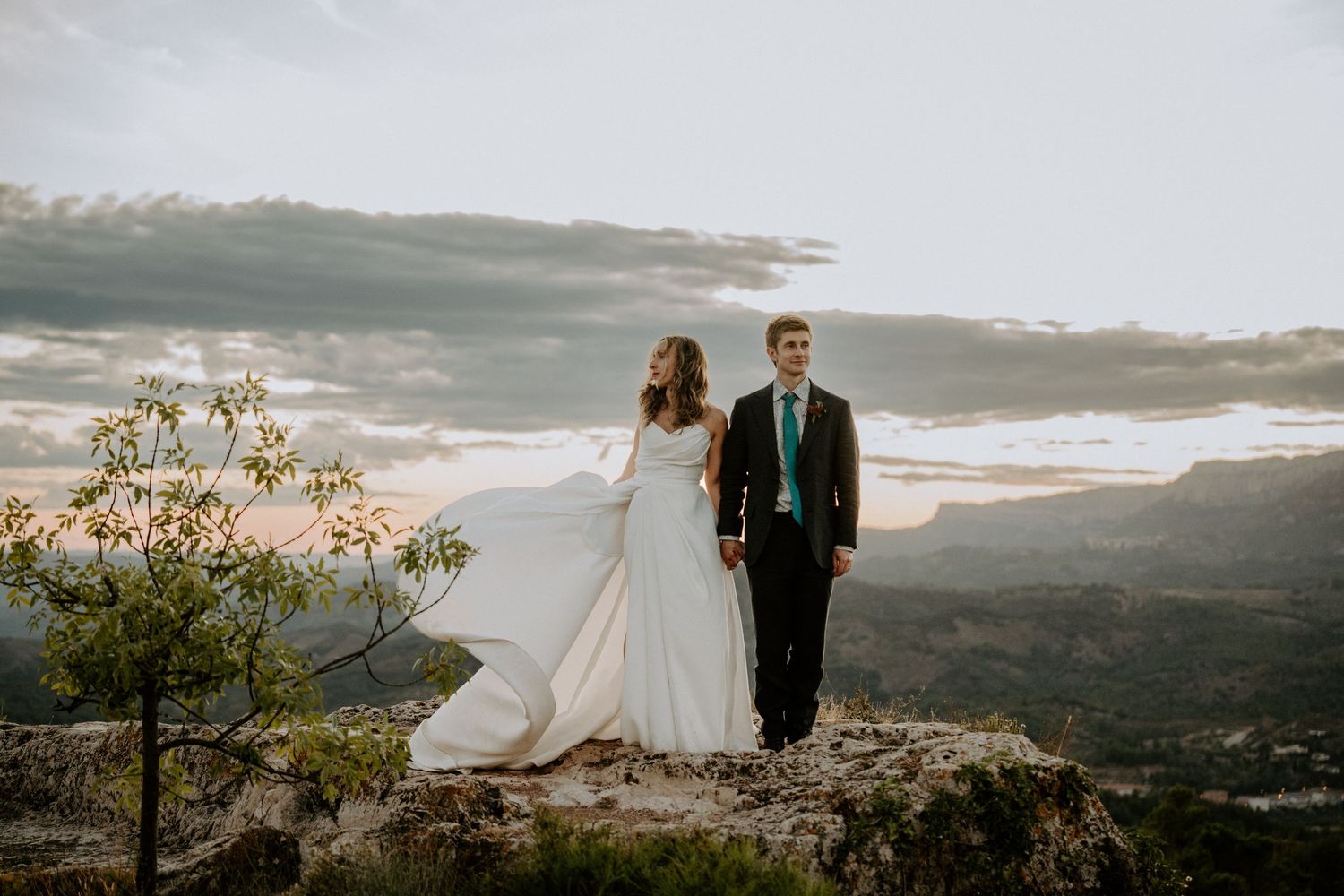 Couple in wedding attire standing on mountain overlook during sunset with dramatic landscape views.