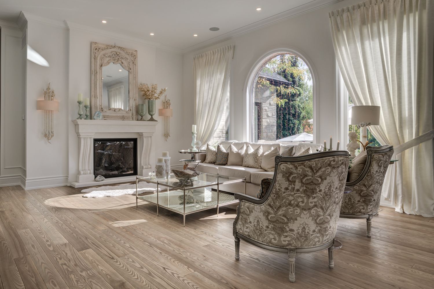 Elegant living room with arched windows, white fireplace, beige sofa, and gray damask chairs on light hardwood floors.