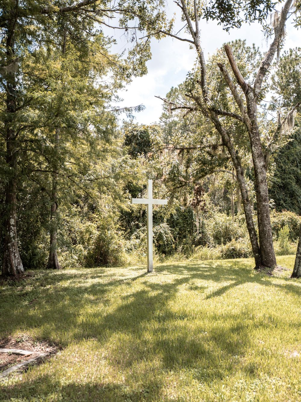 A white cross stands solemnly against a backdrop of sunlit trees and grass in a tranquil outdoor setting.