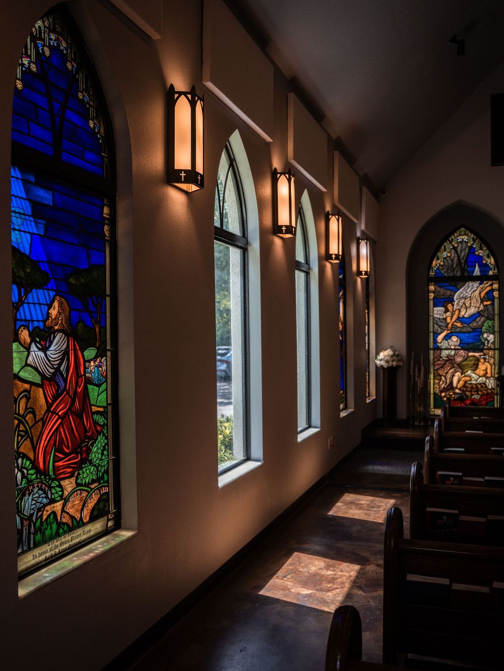 Stained glass church windows cast colorful light along a dark hallway with wooden pews.