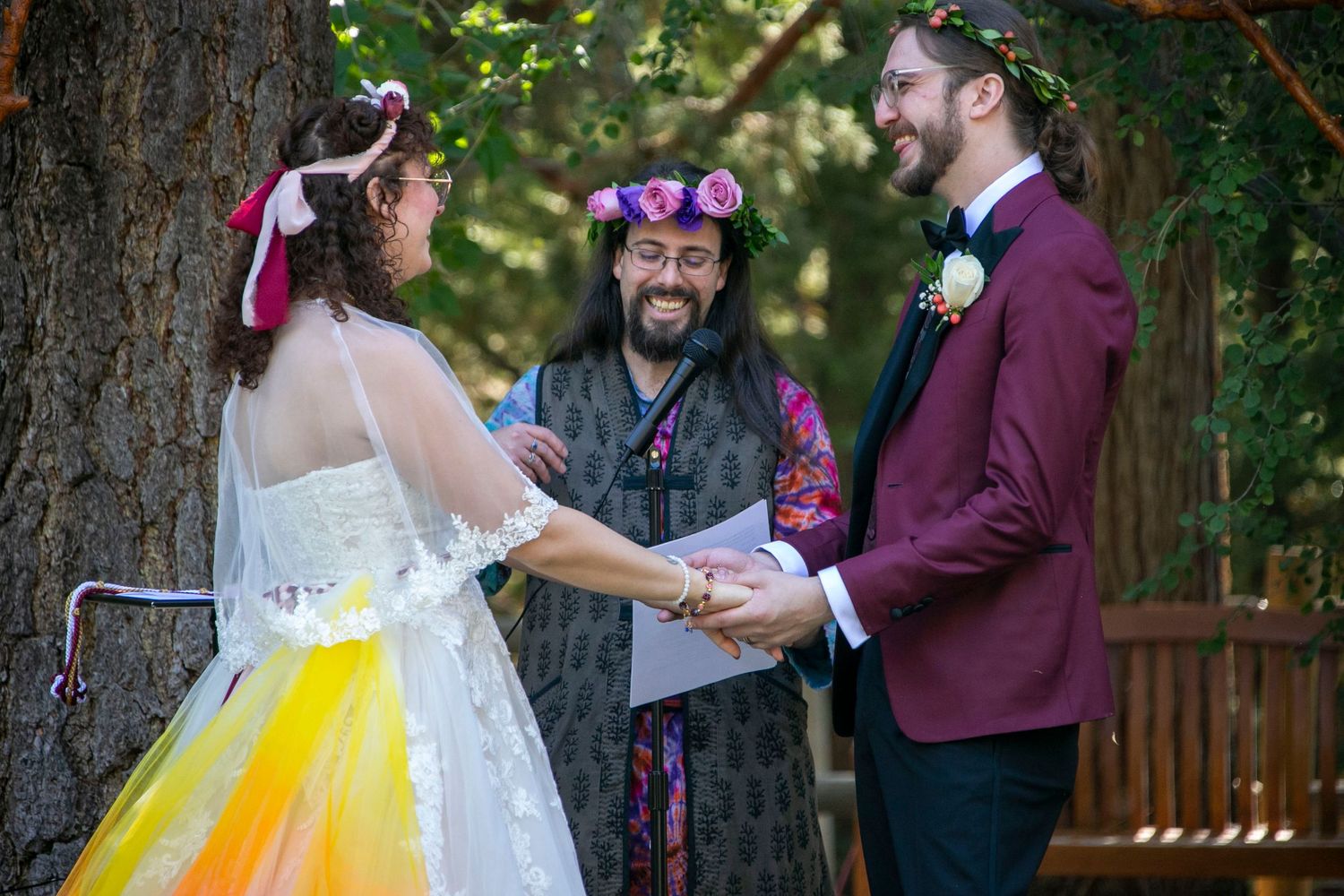 Couple holding hands at outdoor wedding ceremony with officiant, bride in yellow tutu dress, groom in burgundy suit.