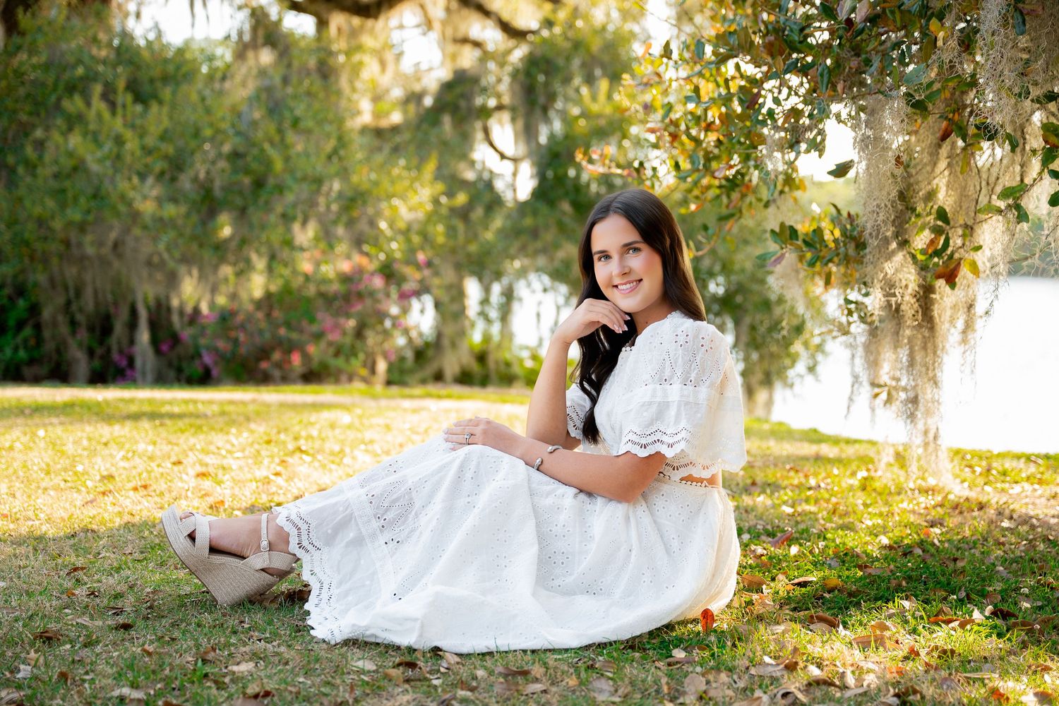Young woman in white dress sitting gracefully on grass under Spanish moss-draped trees in a serene outdoor setting.