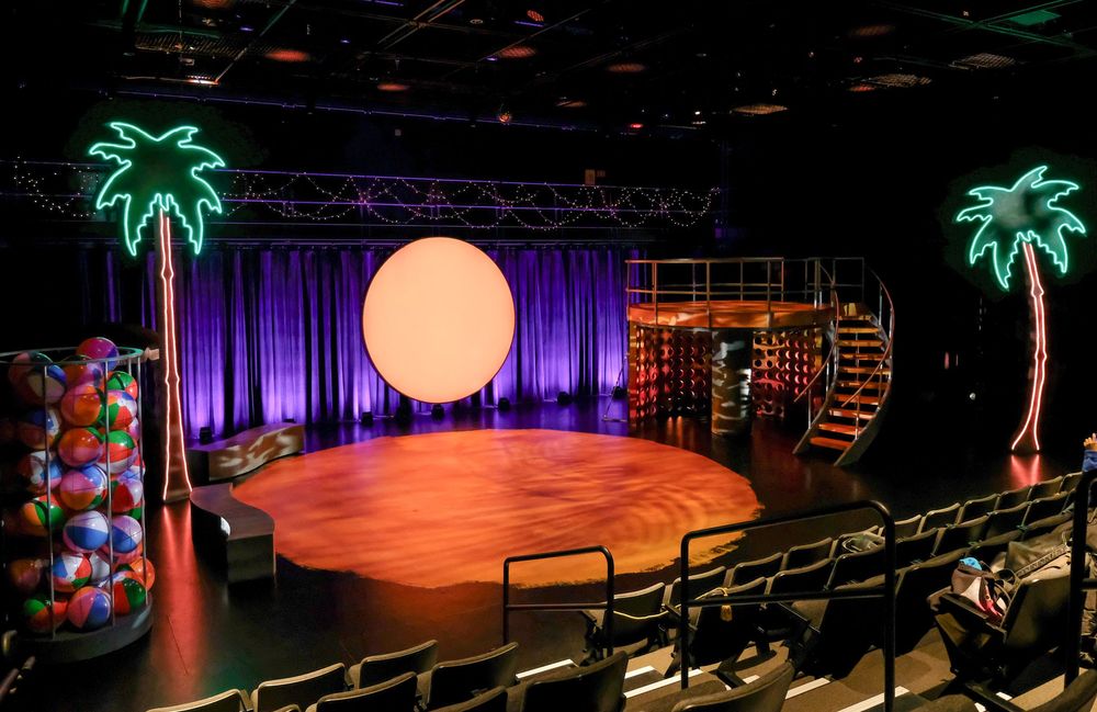 Theater stage with neon palm trees, colorful beach balls, purple curtains, spiral staircase, and glowing moon backdrop.