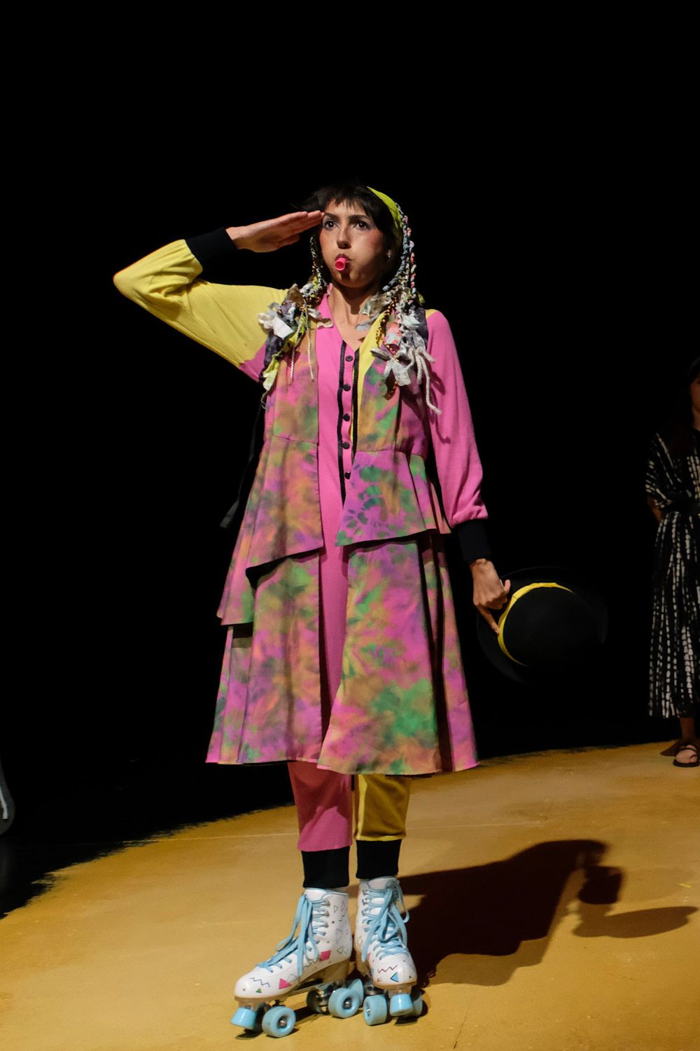 Model in colorful tie-dye outfit with yellow jacket, pink skirt, and white roller skates salutes on runway.