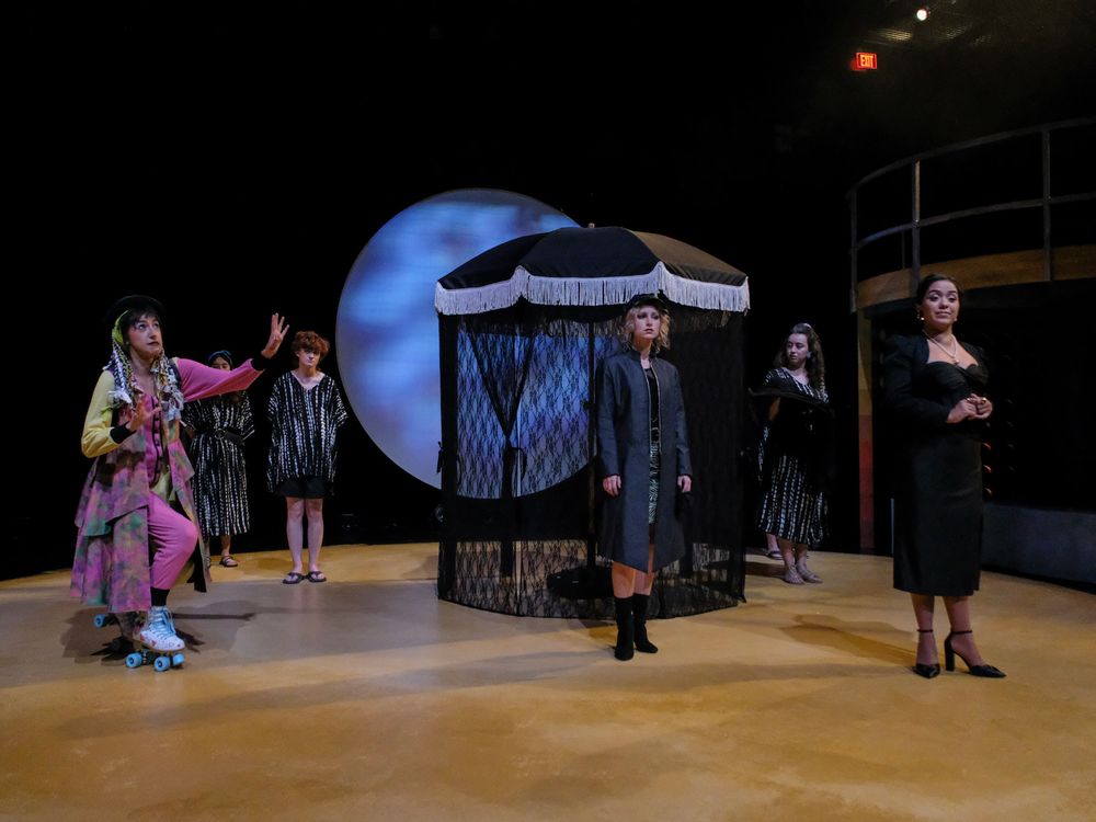 Four performers on a theatrical stage with dramatic black and white costumes under a large moon backdrop.