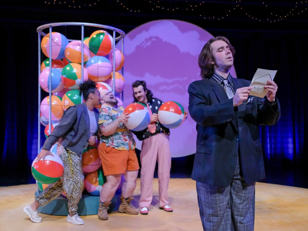 Theater performers on stage with colorful beach balls, purple backdrop, and an actor reading a letter in a playful scene.