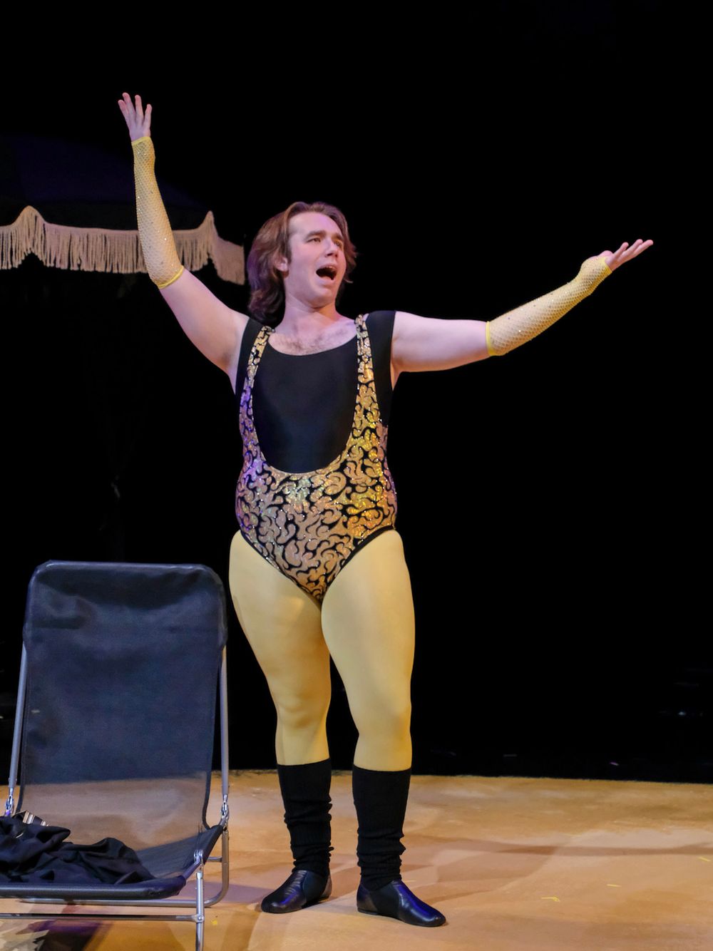 Performer in gold and black leotard with yellow tights and boots, arms raised on stage.