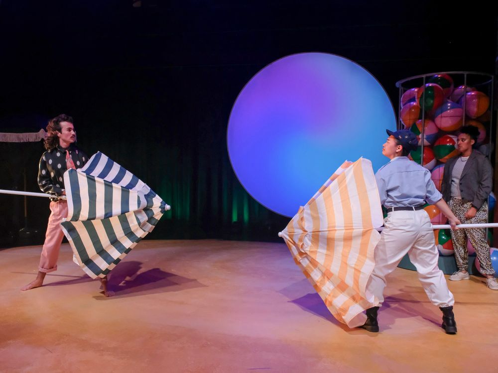 Two performers wave large striped fabric panels on stage with a glowing sphere backdrop in a colorful theatrical production.