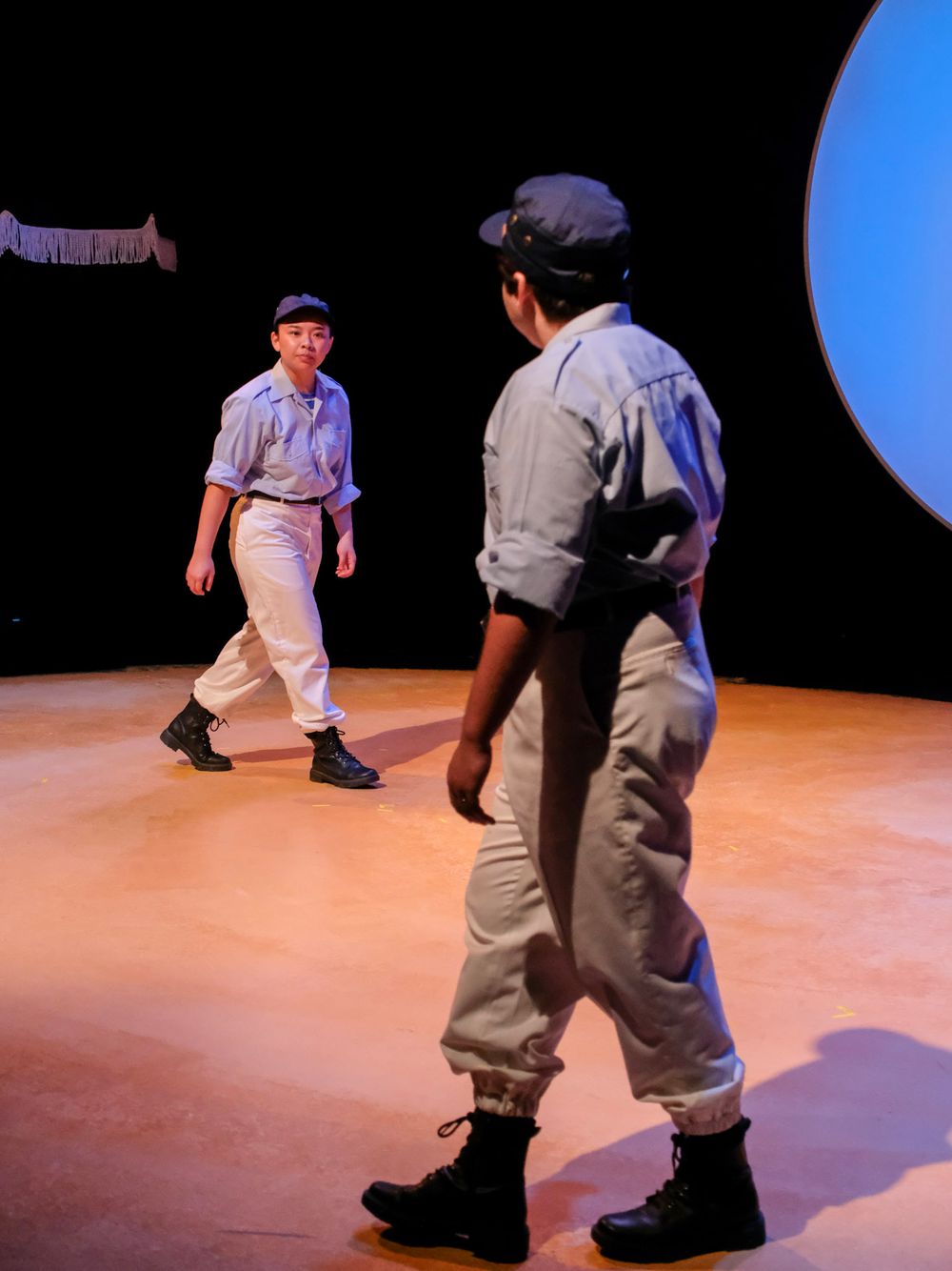Two young performers in matching gray outfits and black boots face each other on a theatrical stage with dramatic lighting.