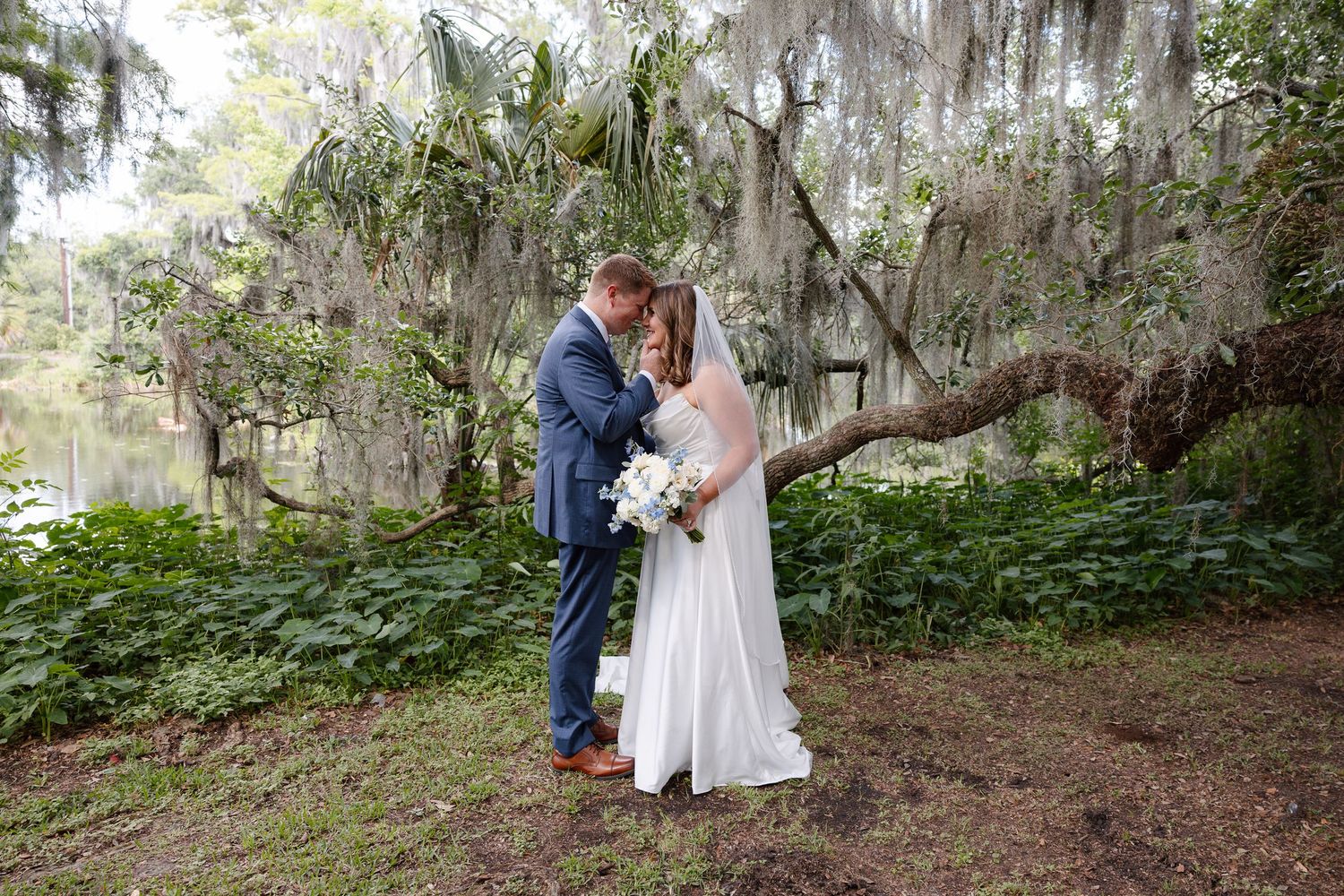 Bride and groom share a romantic kiss under Spanish moss-draped oak trees near a serene pond on their wedding day.